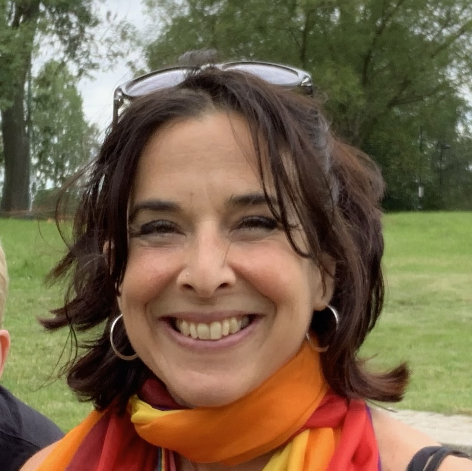 Photo of Harriet wearing sunglasses on her head and a rainbow scarf, smiling and looking directly at the camera, with trees and grass in the background