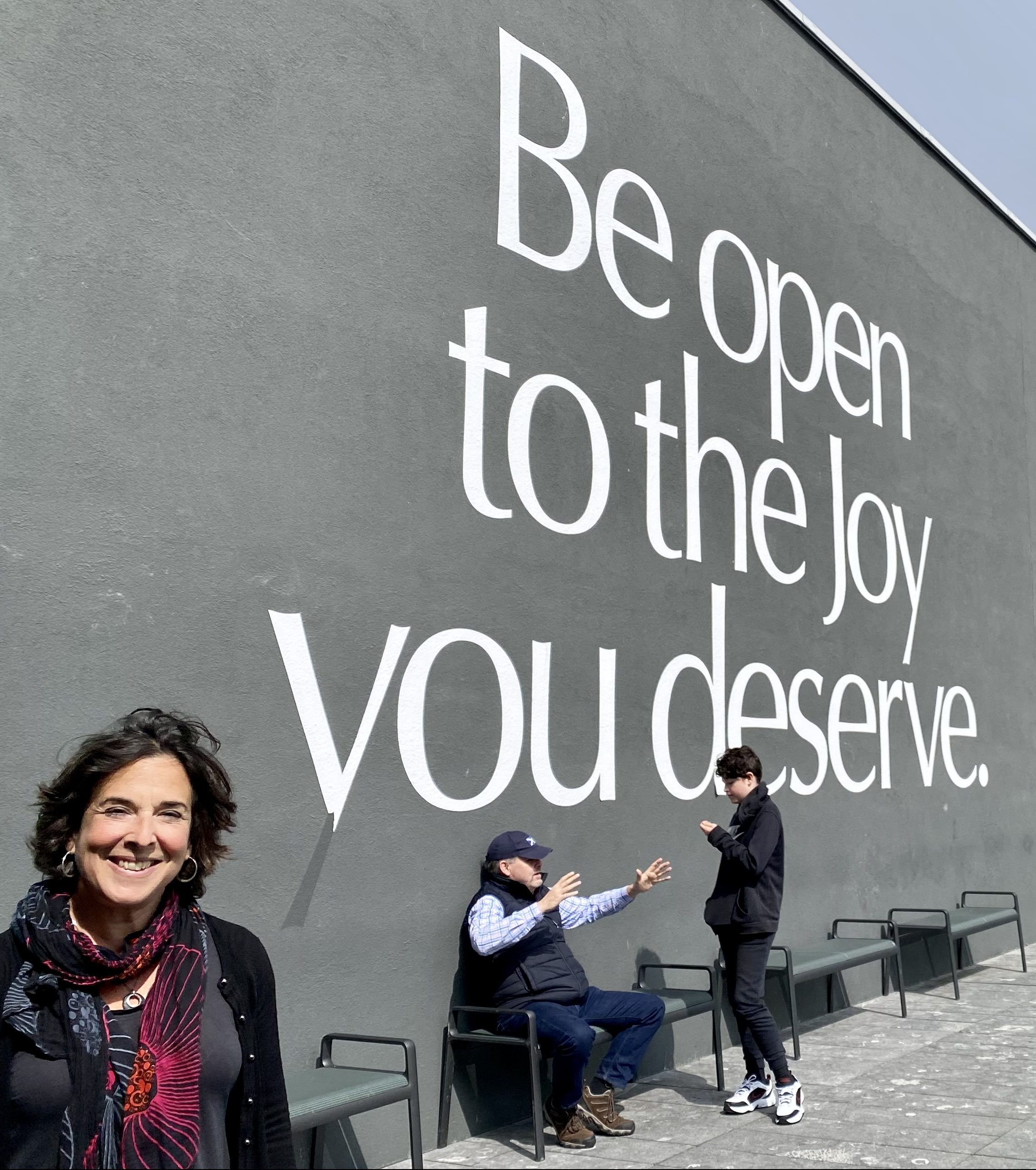 Harriet stands smiling in front of a mural that reads ‘Be open to the joy you deserve’